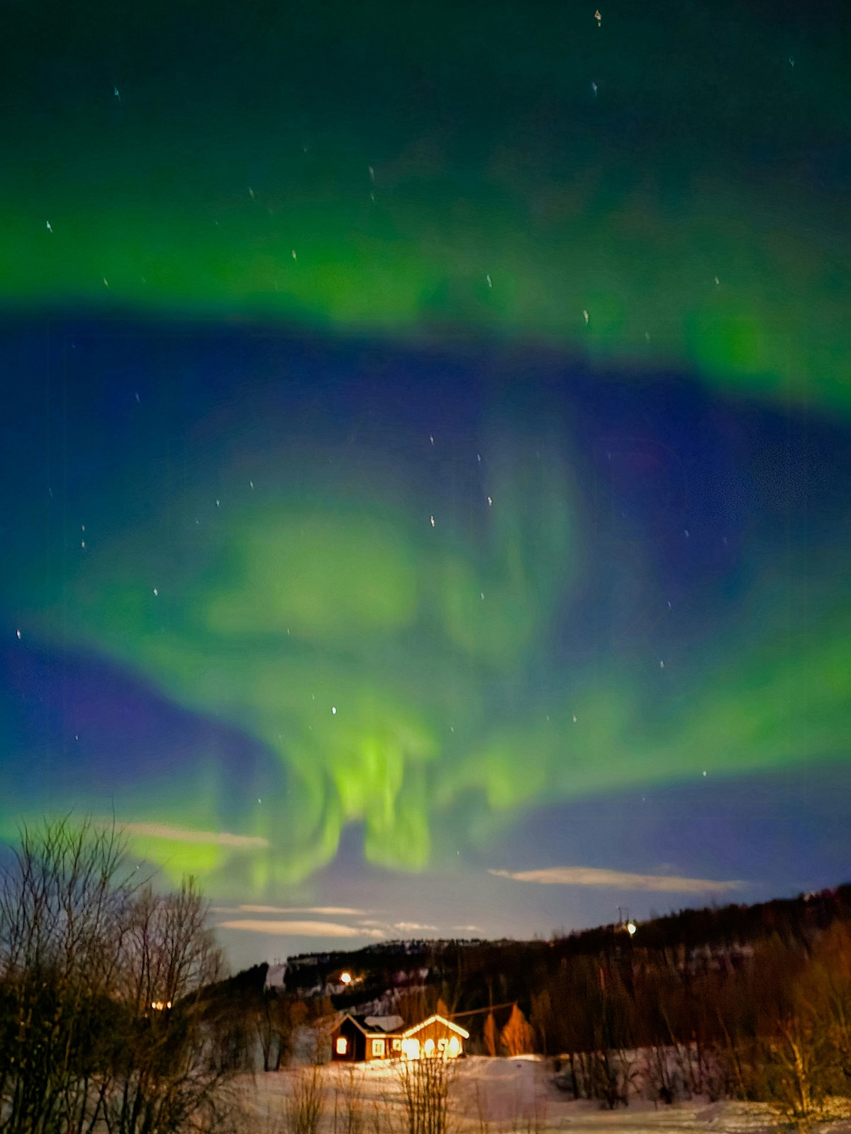 The night sky is illuminated by vibrant green auroras, dancing above a cabin nestled in the landscape. Stars twinkle in the background, and a soft glow emanates from the cabin, contrasting with the surrounding dark terrain and trees.