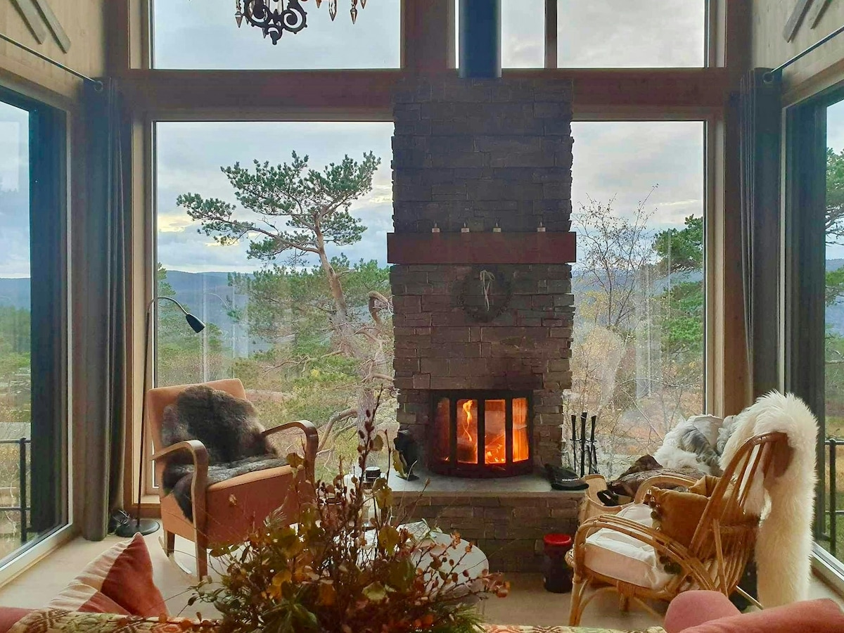 A modern cabin living area features a stone fireplace as its focal point, with a warm fire glowing inside. Large windows provide a scenic view of the surrounding trees and mountains, while comfortable seating invites relaxation. Soft textiles and natural decor elements enhance the cozy atmosphere.