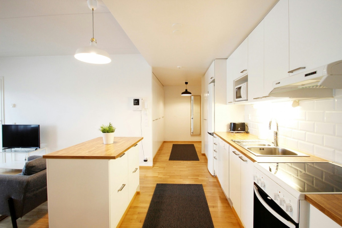 A modern kitchen area features white cabinetry and wood countertops, with an electric stove and microwave. A dining table is situated on the left, and a cozy sitting area is seen in the background. Soft lighting enhances the open and airy layout leading to the hallway.