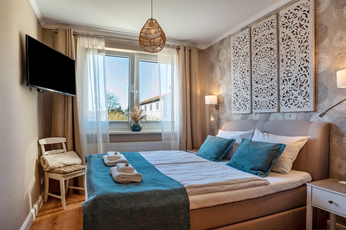 A well-appointed sleeping area is highlighted by a spacious bed dressed in light linens and soft blue accents. Natural light filters through a large window, framed by soft curtains. A decorative wall feature adds visual interest, alongside a wall-mounted television and thoughtfully placed lamps.