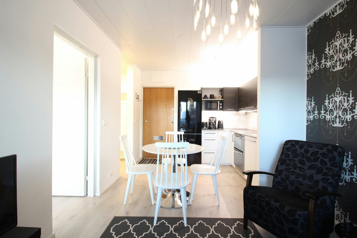 A modern dining area is captured with a round table surrounded by six white chairs, set against a black and white patterned wall. A sleek kitchen is visible in the background, showcasing dark cabinetry and stainless steel appliances. Soft lighting is provided by a unique overhead fixture.