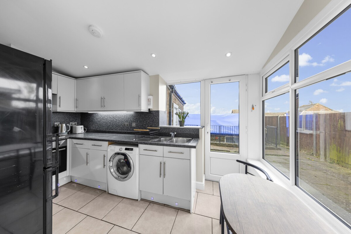 A modern kitchen space is showcased with sleek cabinetry and a dark countertop. Appliances include a washing machine and a kettle. Large windows allow natural light to fill the room, offering views of the outdoor area. A small table is positioned for casual dining.