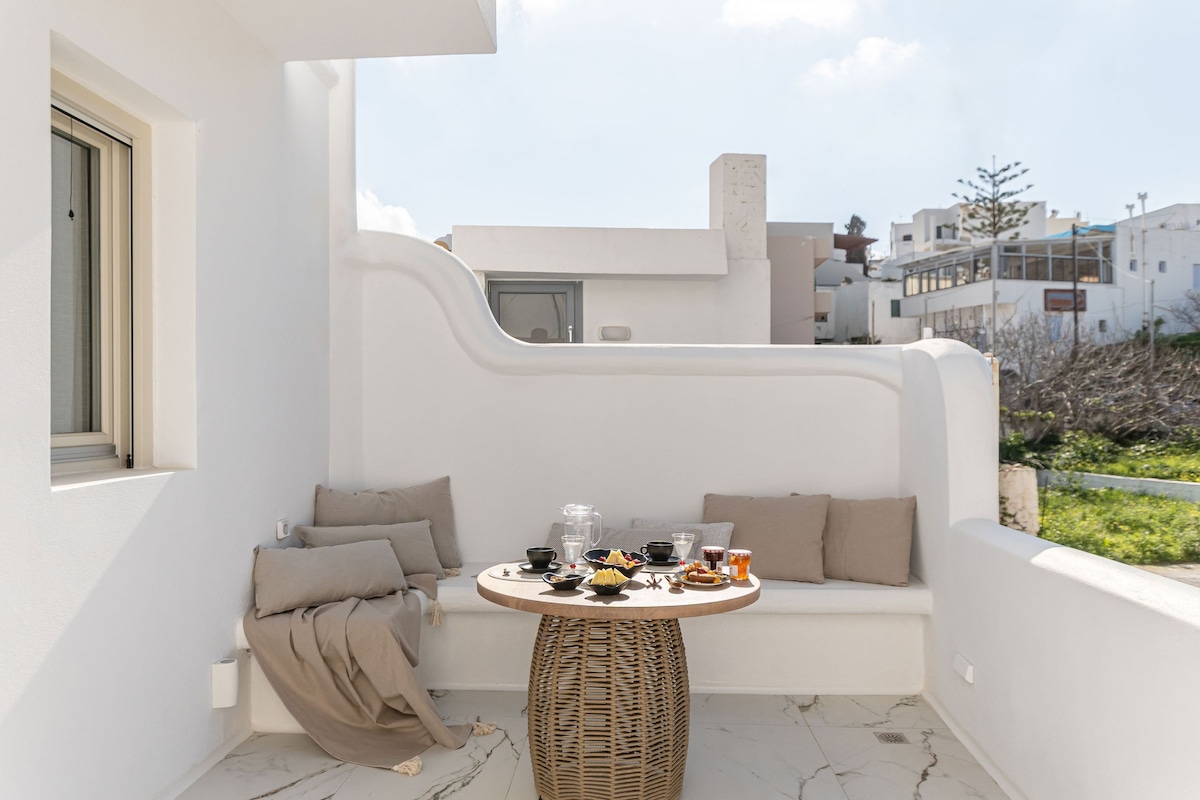 A cozy outdoor seating area is presented with a round table set for a meal, complemented by assorted refreshments and snacks. Soft cushions are arranged against the white walls, providing comfort. Lush greenery is visible in the background, enhancing the serene outdoor space.