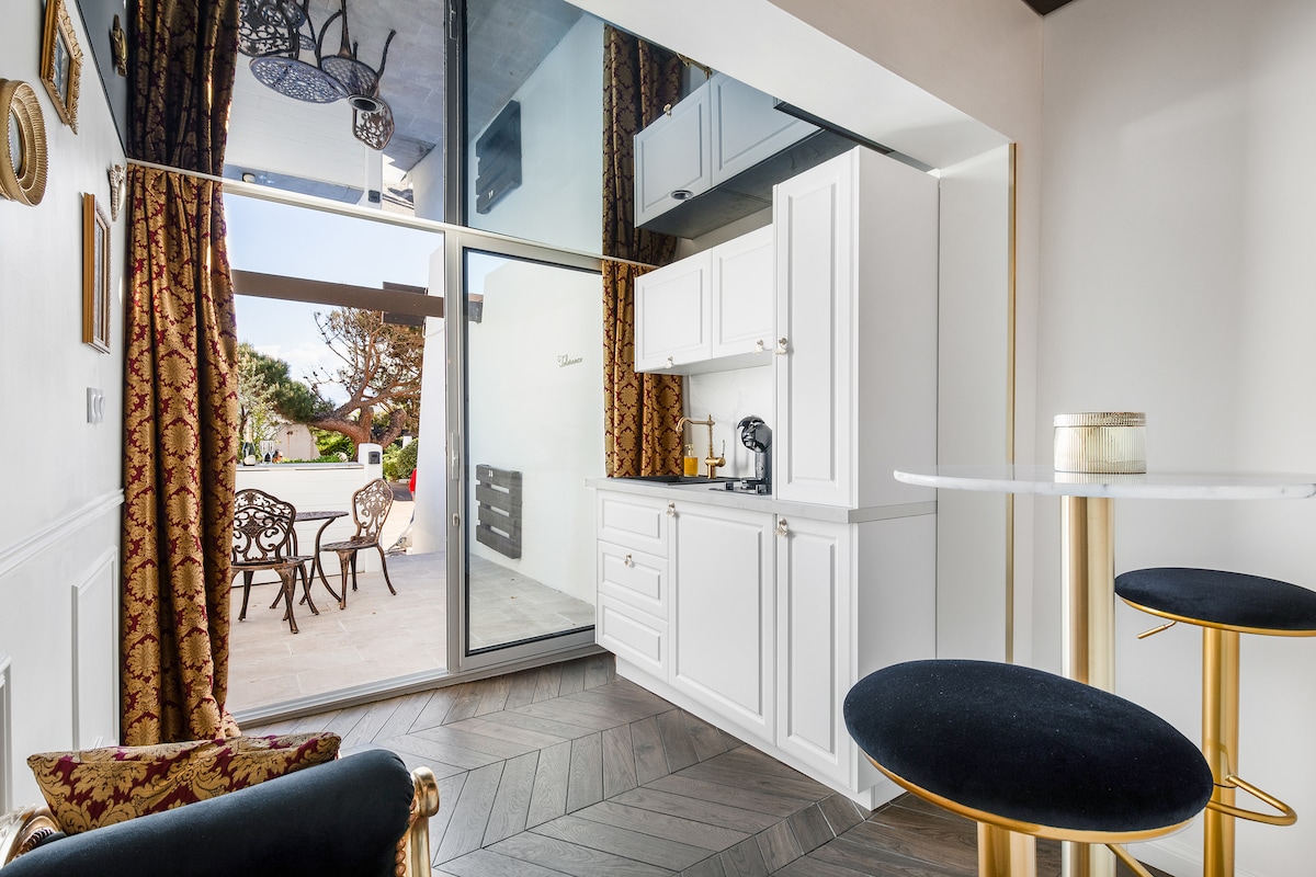 A modern kitchen is visible, featuring sleek cabinetry and a coffee maker. The dining area is represented by a high table with two black stools. Large glass doors open to a terrace, revealing outdoor seating and lush greenery.