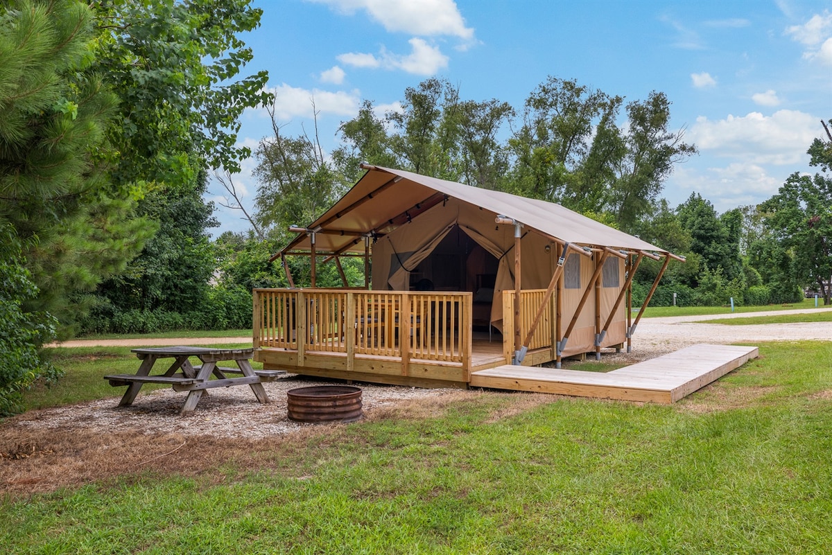 A luxury glamping tent is situated on a grassy area, featuring a wooden deck and a ramp for accessibility. A picnic table is placed nearby, along with a fire pit surrounded by gravel. Green trees and blue skies provide a natural backdrop.