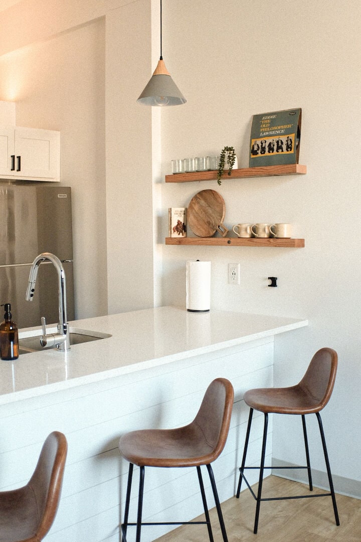 The kitchen features a modern white countertop with three stylish bar stools underneath. A sleek faucet is prominent at the sink, while open shelves display wooden and ceramic items, enhancing the contemporary aesthetic. A stainless steel refrigerator is visible in the background.
