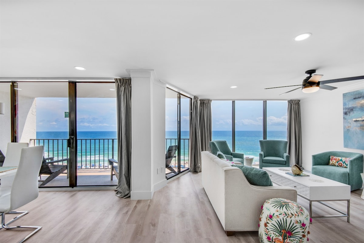 A bright living area features floor-to-ceiling windows offering expansive views of the ocean. Comfortable seating includes a white sofa and teal armchairs, complemented by a stylish table. Nearby, an open dining area is illuminated by natural light flowing through the sliding doors to the balcony.