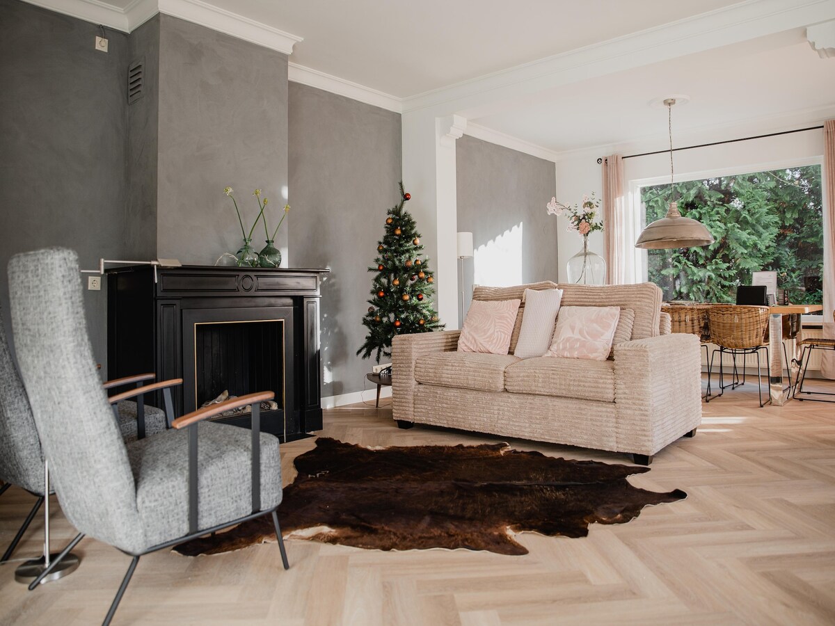 A living area with a textured gray wall and light wooden flooring is shown. A cozy sofa with pillows is positioned near a decorative fireplace, complemented by a stylish armchair. A dining area with a large table and natural greenery is visible through the window.