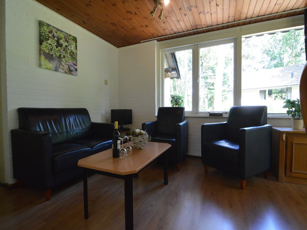 A living area features two black leather armchairs and a matching sofa, arranged around a wooden coffee table. Large windows allow natural light to fill the space, providing views of the surrounding greenery. A small plant sits on the table, enhancing the serene atmosphere.