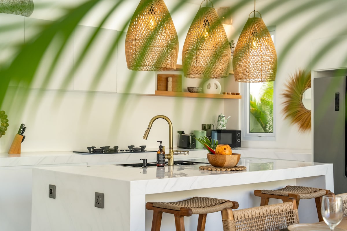 A modern kitchen features a sleek marble countertop with a built-in stove and decorative fruit centerpiece. Three woven pendant lights hang above, providing warm illumination. The space is complemented by natural elements, including nearby greenery visible through a window.