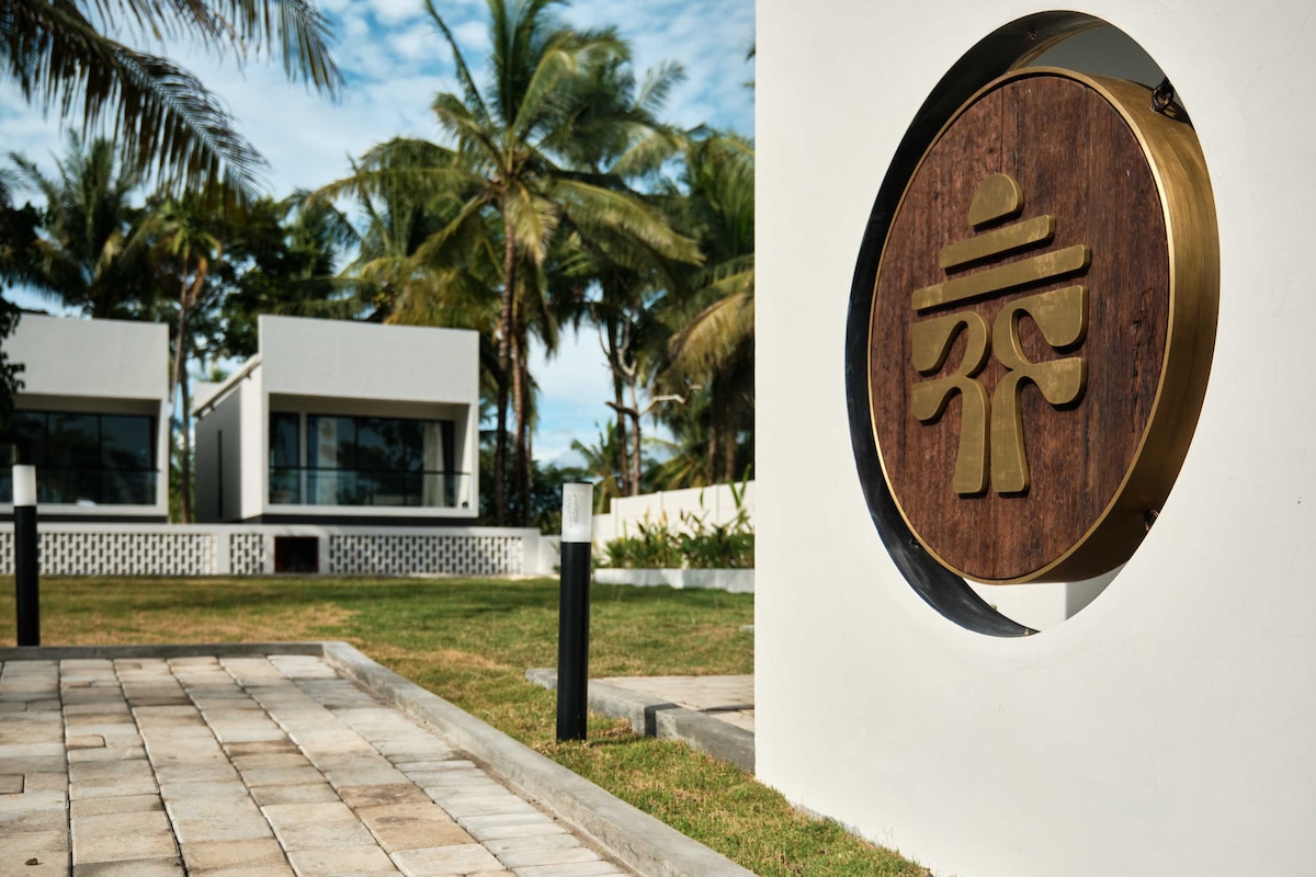 A wooden circular sign featuring a traditional emblem is displayed prominently at the entrance. The surrounding area includes a paved pathway leading to modern villas, framed by lush greenery and palm trees under a clear sky.