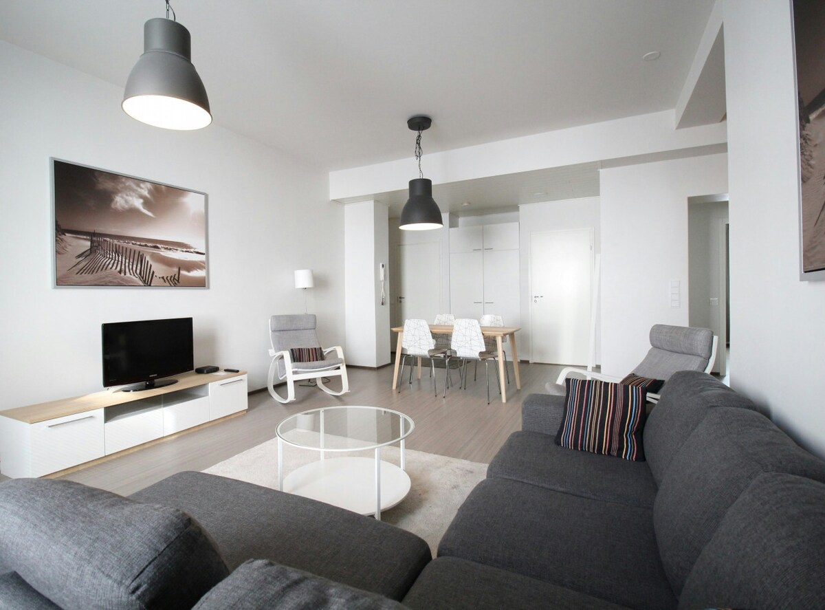 A spacious living room is presented with modern furnishings. A sectional dark gray sofa and a glass coffee table occupy the center, while a flat-screen television is positioned on a low entertainment unit. Two light-colored chairs and a dining table are visible in the background.