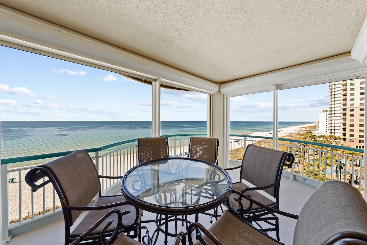 A spacious balcony is furnished with a round glass table surrounded by six metal chairs. The view beyond showcases the expansive Gulf waters and sandy beach, with distant buildings lining the coastline under a clear blue sky.