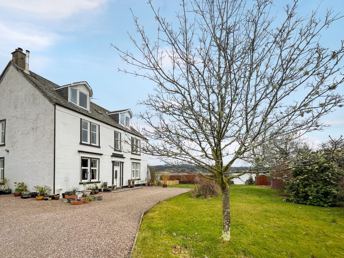 The exterior of Glengilp Apartment is set on a spacious gravel driveway, framed by a well-kept garden with various plants. A leafless tree stands prominently in the foreground, enhancing the welcoming atmosphere of this charming space.