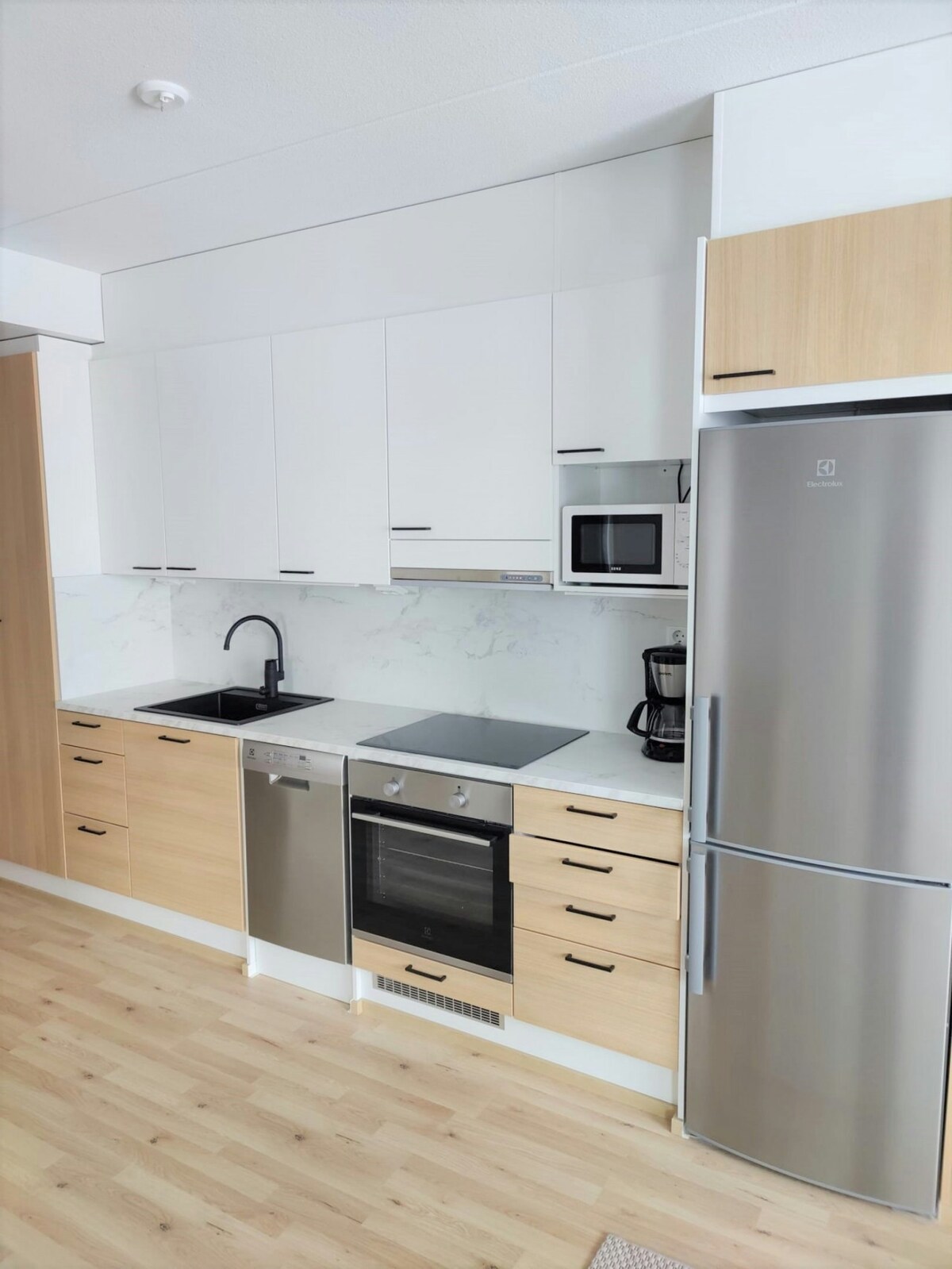 A modern kitchen equipped with sleek appliances. Stainless steel finishes include an oven, microwave, and refrigerator, complemented by a coffee maker. Wooden cabinetry offers storage, and a black sink is integrated into the countertop, providing a clean and functional workspace.