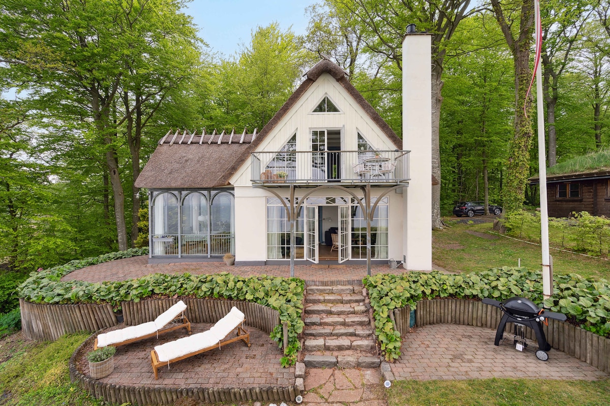 A charming thatched cottage stands amidst lush greenery, featuring multiple large windows that allow natural light to flood the interiors. Sun loungers are positioned on a terrace, while a gas grill is ready for outdoor cooking. The stone pathway leads to an inviting entrance.