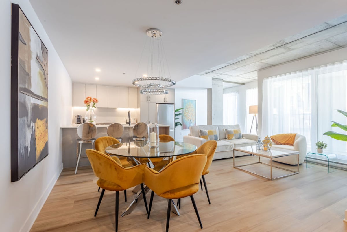 A modern living area is showcased with a round glass dining table surrounded by vibrant yellow chairs. A comfortable sofa sits nearby, complemented by natural light filtering through sheer draperies. A contemporary chandelier hangs above, adding to the inviting atmosphere of the open space.