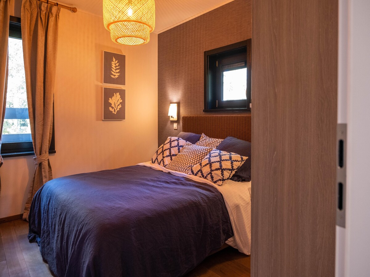 A spacious bedroom is featured with a large bed dressed in a deep blue coverlet. Textured walls are accented by framed botanical prints. Natural light filters through a window, and soft drapes provide an inviting touch. A warm light fixture offers a cozy ambiance.