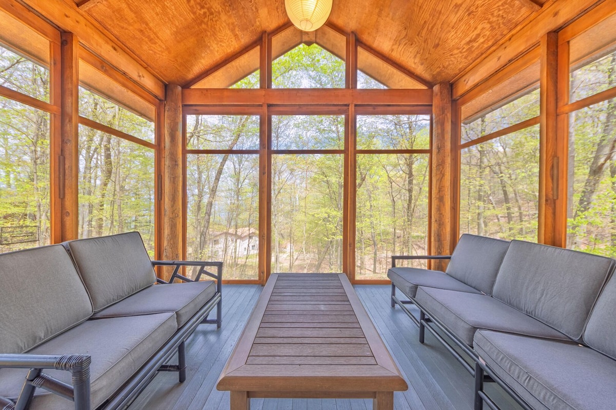 A screened-in porch features large windows that allow natural light to fill the space, surrounded by trees. Two comfortable couches are positioned around a wooden coffee table, creating a relaxing area for guests to enjoy the serene outdoor views.