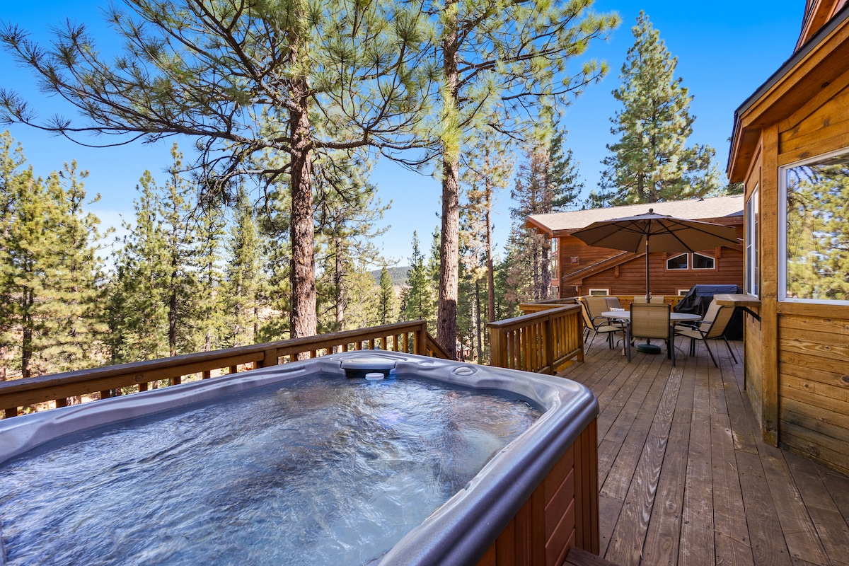 A private deck features a hot tub surrounded by trees, offering tranquil views of the natural landscape. Comfortable seating arrangements are positioned nearby under an umbrella, creating an inviting space for relaxation and enjoying the outdoors.