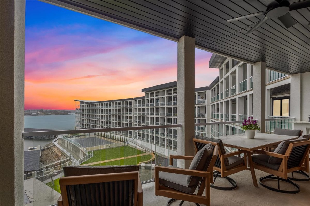 A spacious balcony offers stunning views of a vibrant sunset over the lake. Comfortable seating is arranged around a dining table, set against the backdrop of the modern building and well-maintained grounds below.