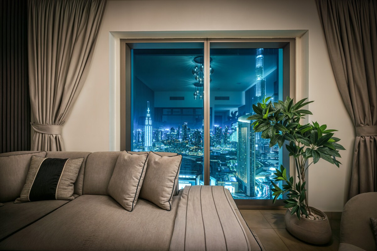 A cozy seating area featuring a comfortable sofa is positioned near a large window, offering stunning views of the Dubai skyline at night. Soft curtains frame the window, while a decorative plant adds a touch of greenery to the space.