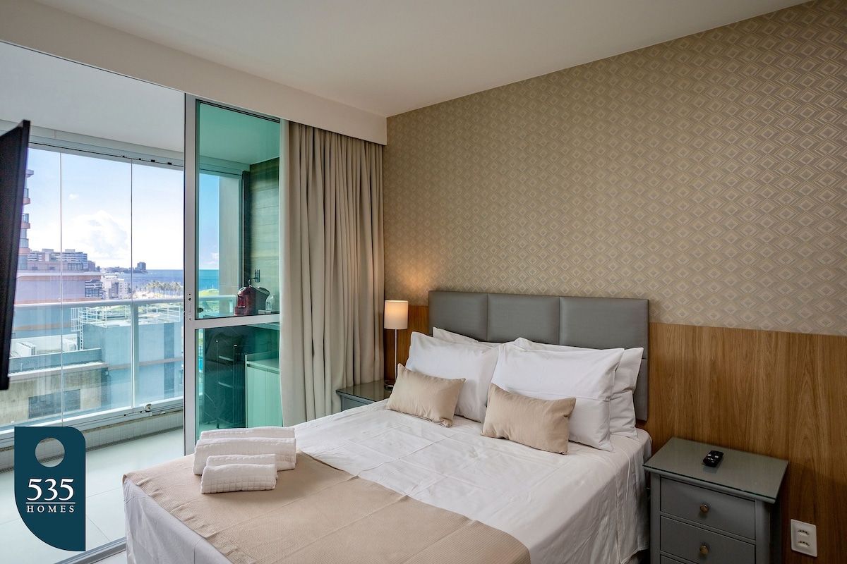 A well-appointed bedroom features a spacious bed with neatly arranged pillows. Natural light streams through floor-to-ceiling windows, offering glimpses of the ocean view. Soft, neutral tones create a calming atmosphere, complemented by wooden accents and bedside tables.