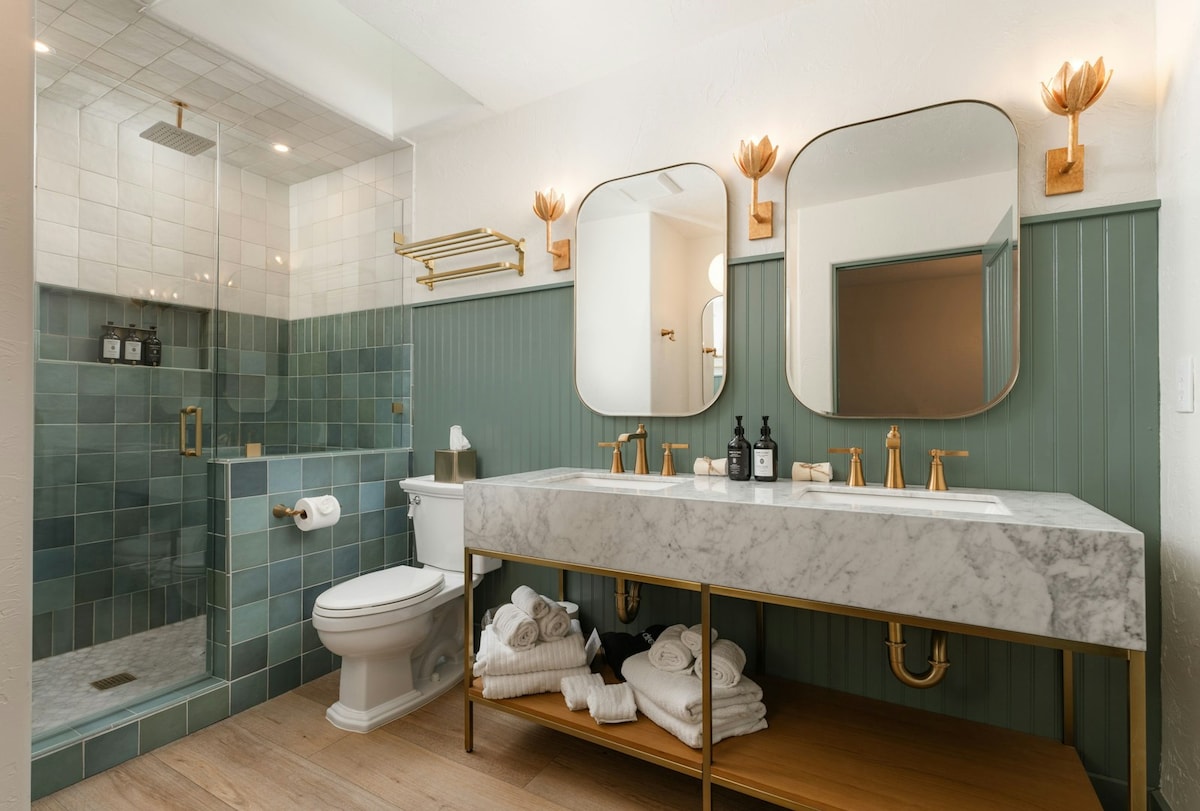 The spacious bathroom features a walk-in tiled shower with glass enclosure and a double vanity sink. A marble countertop is supported by elegant gold legs, with organized amenities displayed. Soft white towels are neatly arranged on a shelf below. Decorative lighting accents the wall, complementing the soothing color palette.