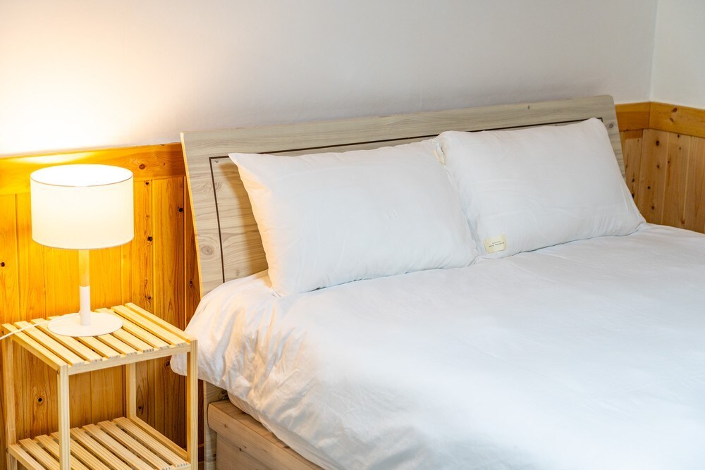 A spacious bed with crisp white linens is positioned next to a wooden nightstand. A soft glow is provided by a bedside lamp, enhancing the serene ambiance of the room. The walls feature a light wood finish, contributing to a cozy atmosphere.