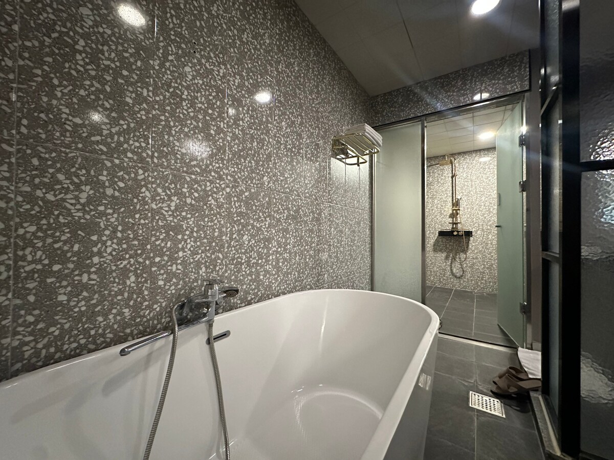 A modern bathroom is featured, showcasing a freestanding bathtub with sleek fixtures. The walls are adorned with textured tiles in shades of gray, while the spacious shower area is enclosed by glass doors, enhancing the open feel of the space.