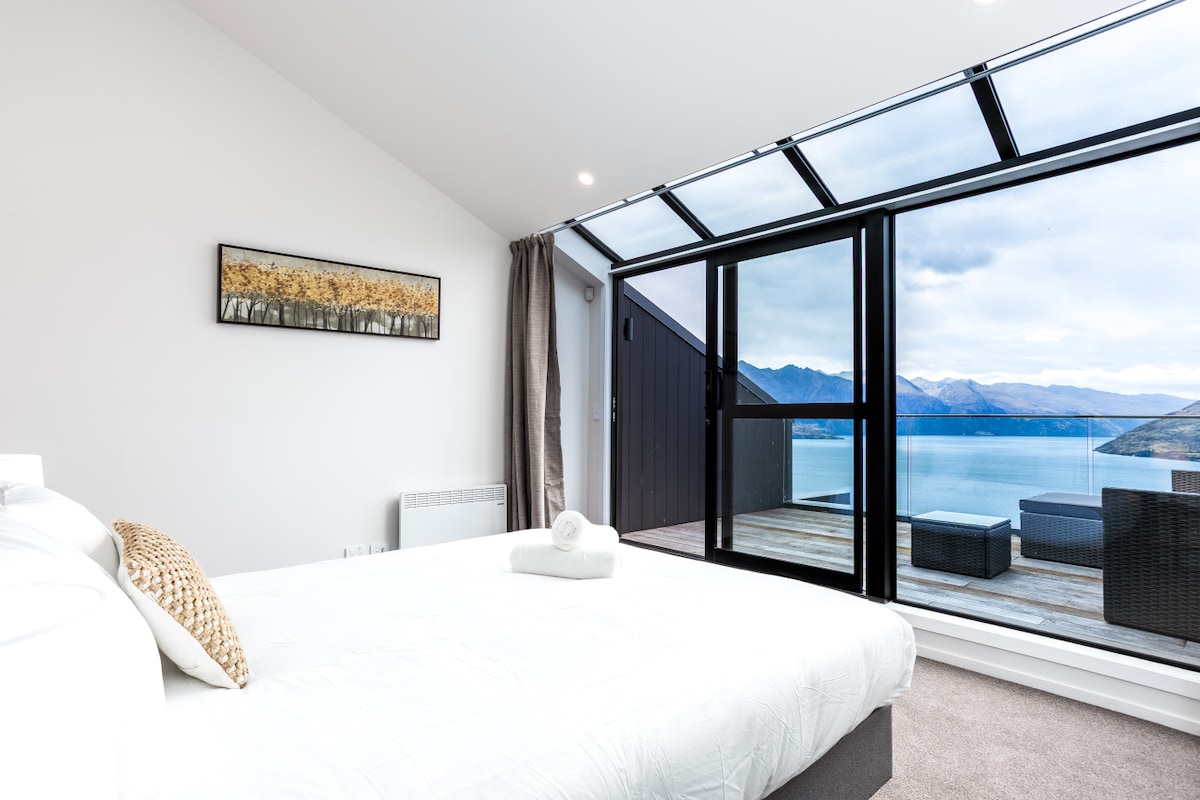 A spacious bedroom features a large bed with white linens and a decorative pillow. Floor-to-ceiling glass doors open to a private balcony offering expansive lake and mountain views. A soft color palette is complemented by natural light flowing into the room.