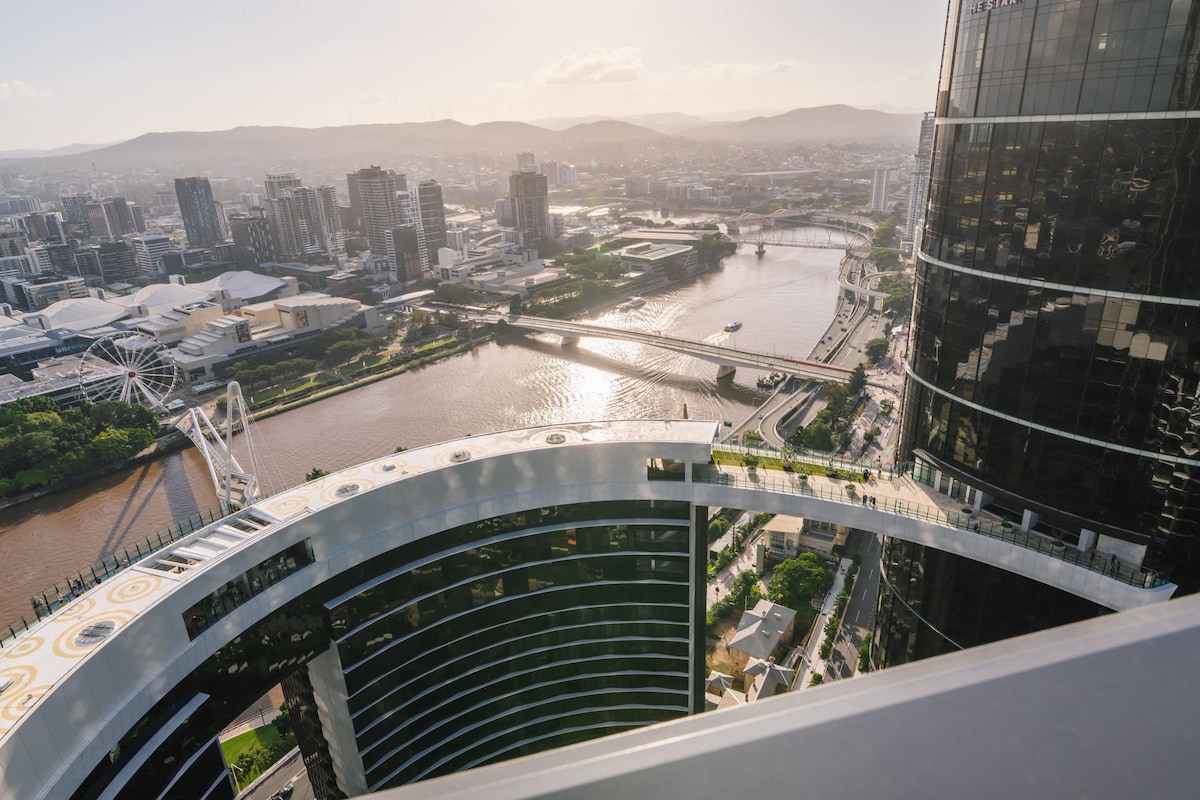 A panoramic city view is captured from a high vantage point, showcasing Brisbane's skyline and the winding river below. The horizon is bathed in a warm morning light, revealing a mix of modern buildings and greenery, with distant hills framing the scene.