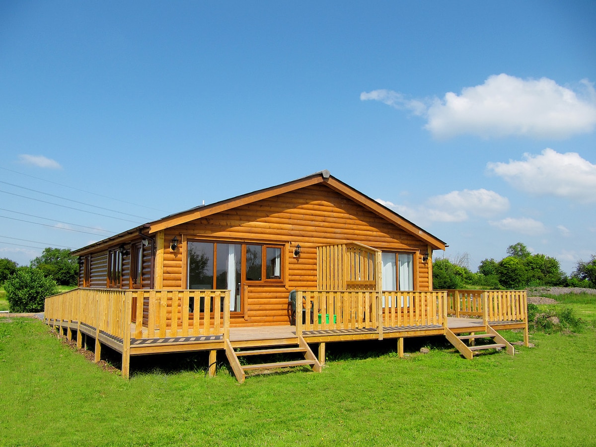 A spacious lodge constructed from timber is depicted, with a large wooden deck surrounding its perimeter. Large windows allow for natural light, and outdoor seating is visible on the deck. A grassy area extends in front of the lodge, under a clear blue sky.