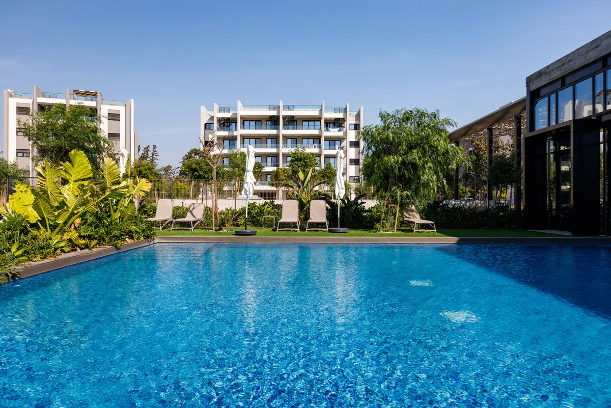 A serene pool area is framed by lush greenery and lounge chairs, inviting relaxation. The bright blue water reflects the clear sky, while a modern building stands in the background, showcasing multiple balconies. Natural landscaping enhances the tranquil setting.