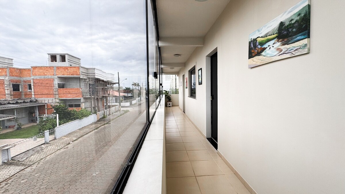 A well-lit corridor features large windows on one side, allowing natural light to fill the space. The walls are painted in neutral tones, with a piece of artwork adding color. Smooth tile flooring extends throughout, providing easy mobility.