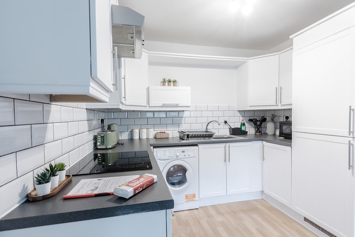 A modern kitchen features sleek white cabinetry and a dark countertop. Essential appliances, including a washing machine and a stovetop, are visible. Subtle greenery is present on the counter, and a bright window provides natural light, enhancing the functional workspace.