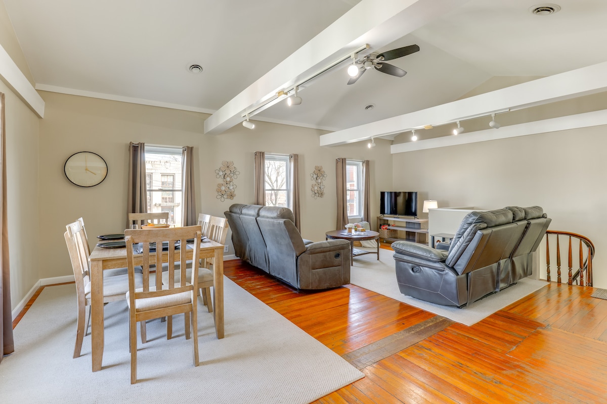 An open living space features a dining area with a wooden table and six chairs. Comfortable leather seating is arranged around a coffee table, facing a TV setup. Large windows allow natural light to illuminate the room, complemented by wooden flooring and a neutral color palette.