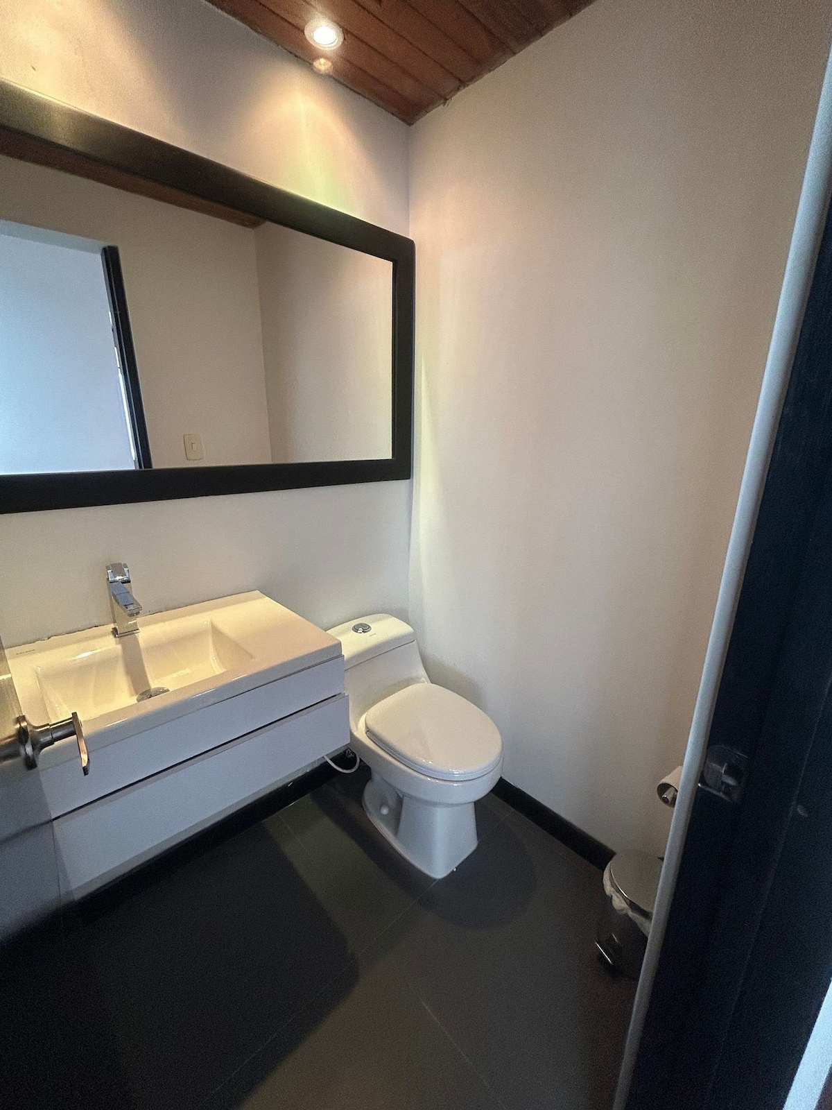 A modern bathroom is shown, featuring a sleek white vanity with a minimalist sink. A large mirror enhances the sense of space, while a clean toilet is positioned nearby. The floor is finished in dark tiles, complementing the overall neutral color palette.