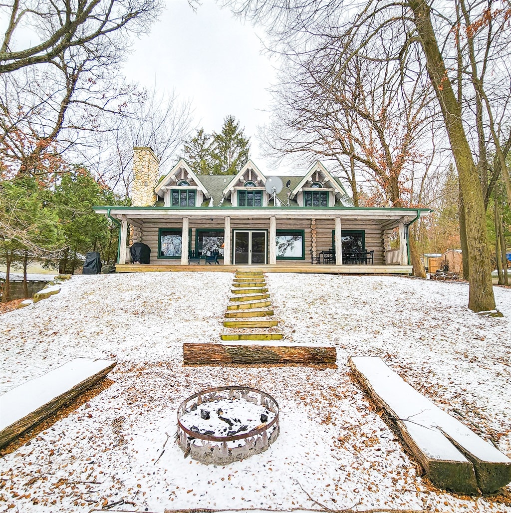 A cabin is set on a snowy sloped landscape, featuring a large deck with multiple seating options. Stone steps lead to the entrance, while a circular fire pit is surrounded by snow. Trees frame the scene, lending a sense of seclusion.
