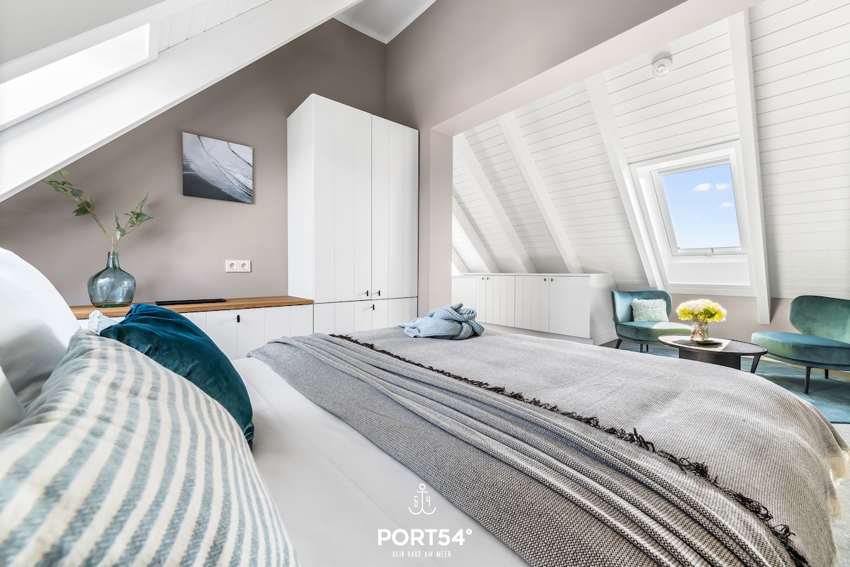 A bright and airy sleeping area features slanted ceilings with skylights, allowing natural light to filter in. A spacious bed is dressed with layered textiles. Soft green chairs are positioned near a small table, while a decorative vase rests on a side unit, enhancing the serene atmosphere.