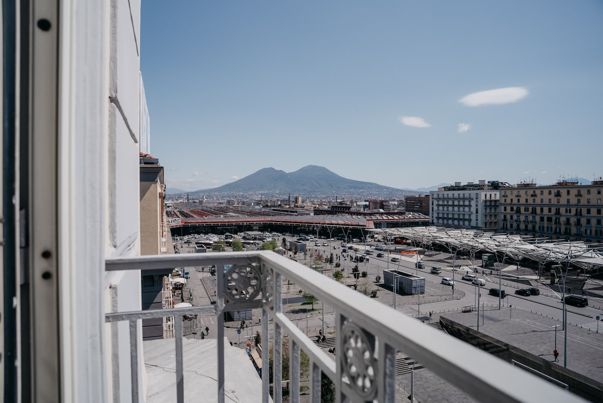 A scenic view of the surrounding landscape is presented from the balcony. The iconic Mount Vesuvius is visible in the background, contrasting with the cityscape below. The nearby train station and urban environment are observed, with clear blue skies and minimal cloud cover enhancing the vista.