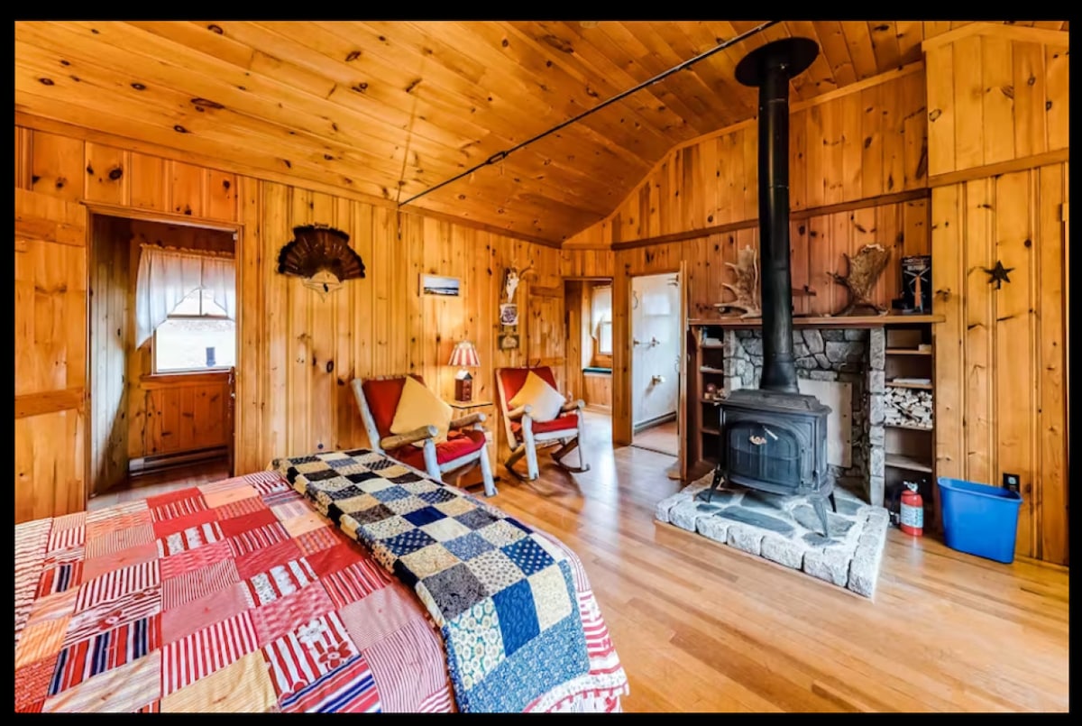The open living space features warm wood paneling and a cozy wood-burning stove positioned against a stone backdrop. A spacious bed draped in a colorful patchwork quilt is complemented by two inviting armchairs. Natural light streams through the nearby window, enhancing the rustic charm.