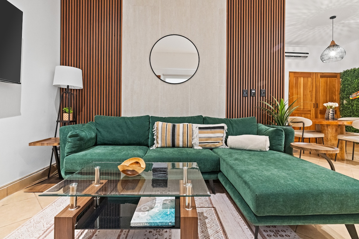 A comfortable living area features a large green sectional sofa, adorned with decorative pillows, positioned against a textured wall. A low glass coffee table sits in front, complemented by a decorative bowl. The space is brightened by natural light, enhancing a welcoming feel.