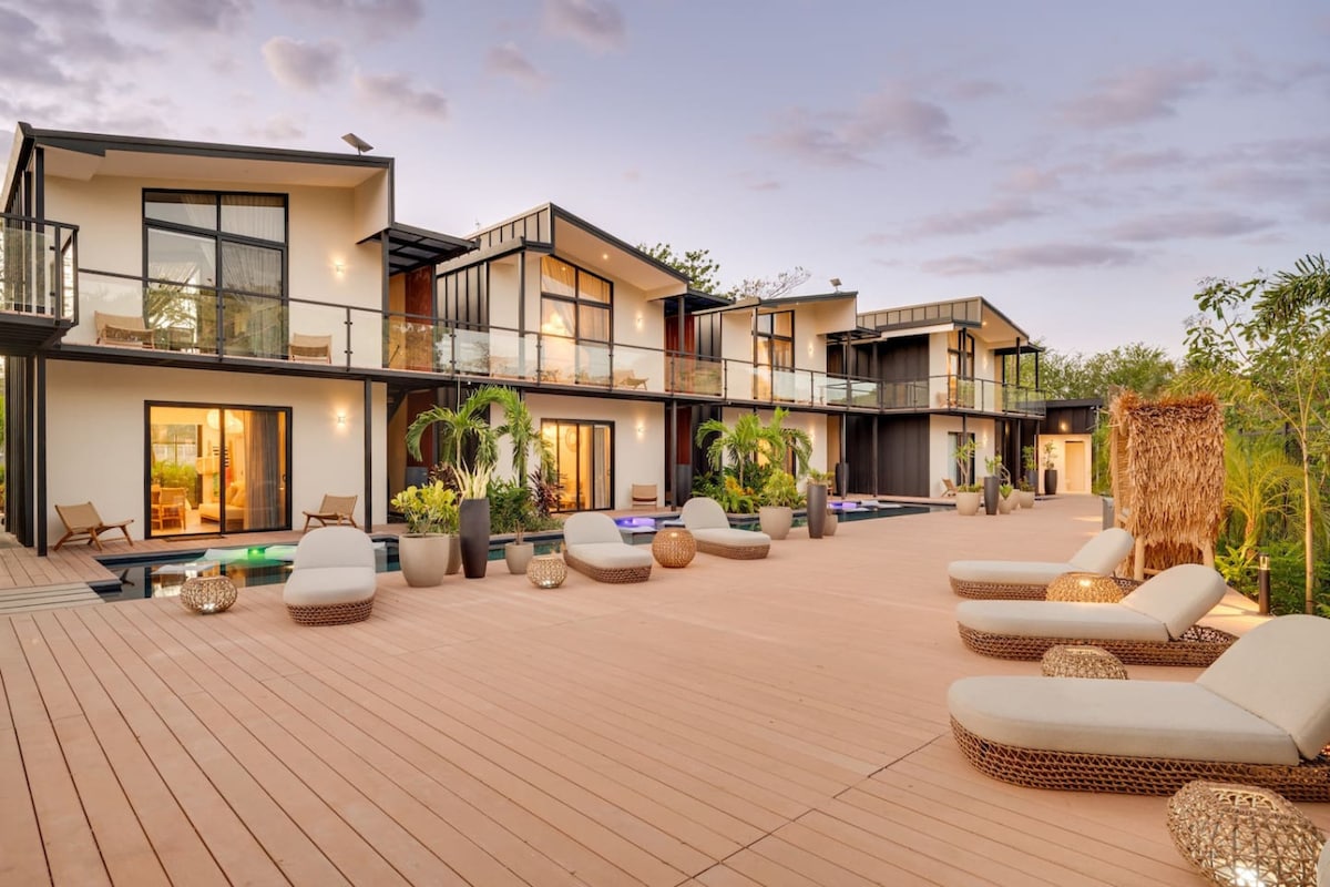 The outdoor area is highlighted by a spacious deck featuring multiple lounge chairs arranged for relaxation. Two distinct buildings are visible, each with sliding glass doors that open to personal balconies. Lush greenery surrounds the property, enhancing the tropical atmosphere.