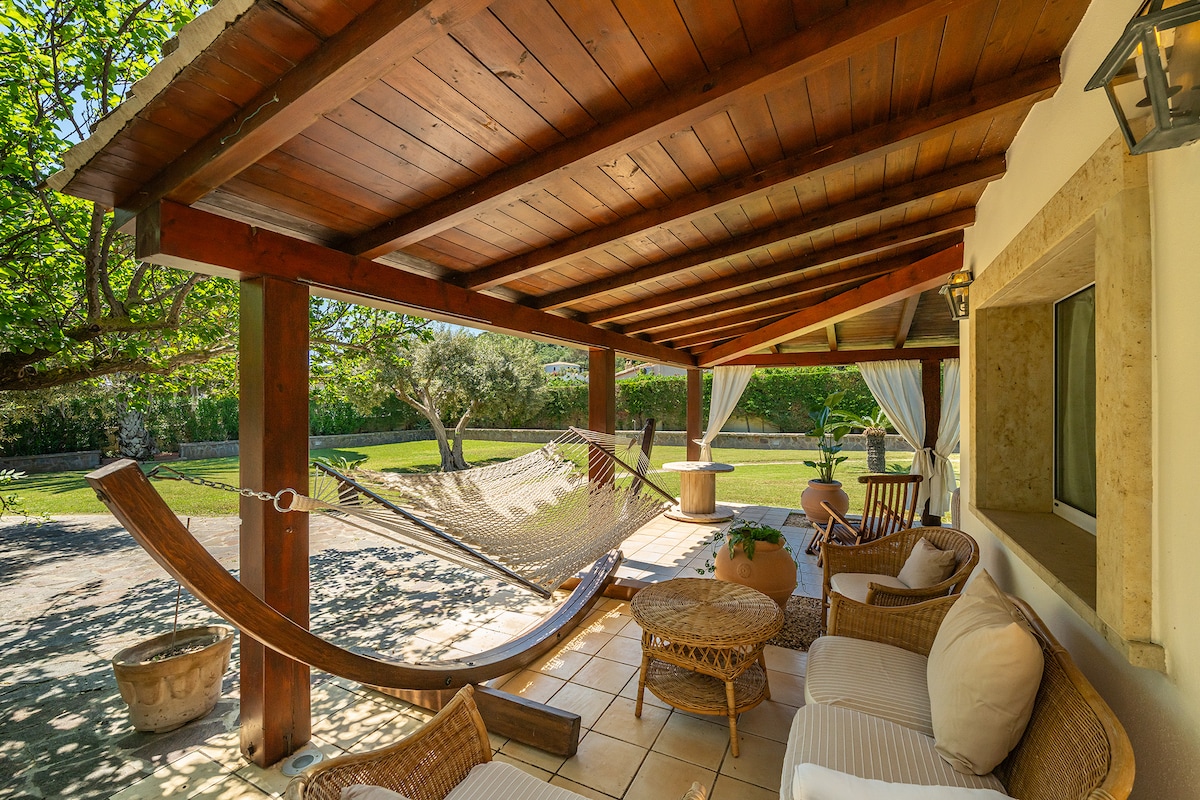 An outdoor covered patio features a hammock and a seating area with wicker sofas and a round coffee table. Natural light filters through the surrounding greenery, creating a peaceful atmosphere, with a spacious garden visible in the background.