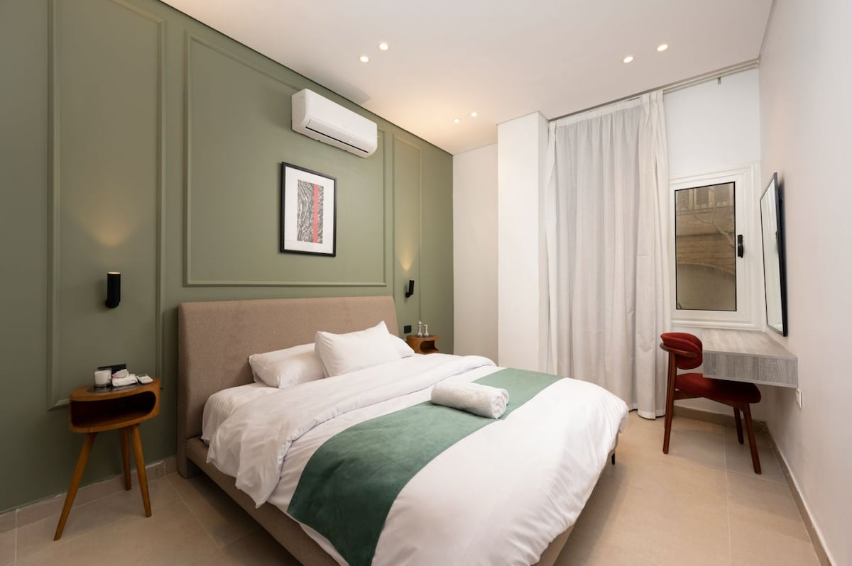 A tranquil sleeping area features a spacious bed with a white duvet and a green accent blanket. A minimalist desk and chair are positioned beside a window with light curtains, while wall-mounted lights provide soft illumination. The calming green walls enhance the serene environment.