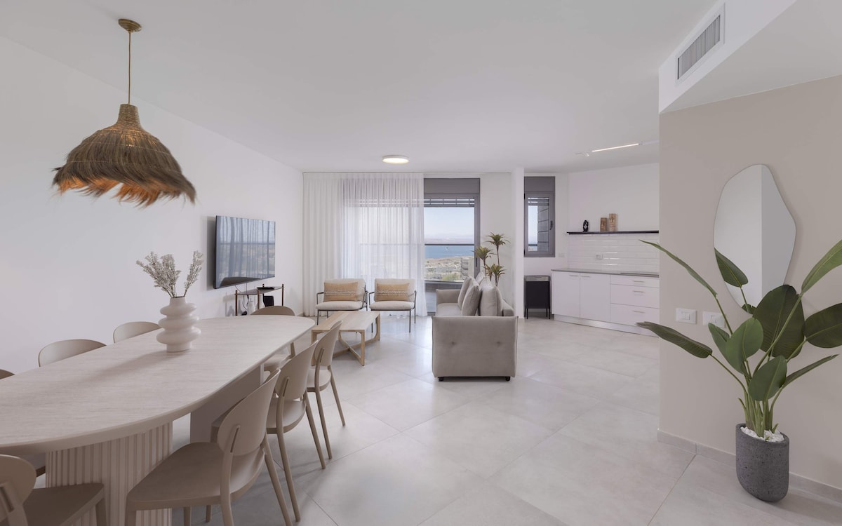 A spacious living area is highlighted by a large dining table surrounded by light chairs. Soft seating is provided by a sofa and two armchairs. Natural light floods the room through large windows, offering views of the surrounding landscape and sea.