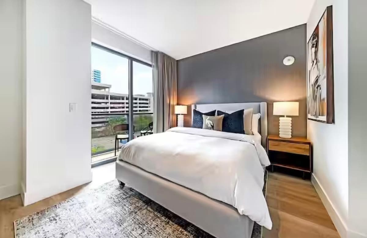 A softly lit bedroom features a spacious bed with a light-colored comforter. Two bedside lamps provide gentle illumination. Large glass doors lead to an outdoor view, complemented by neutral-toned walls and a decorative area rug underfoot.