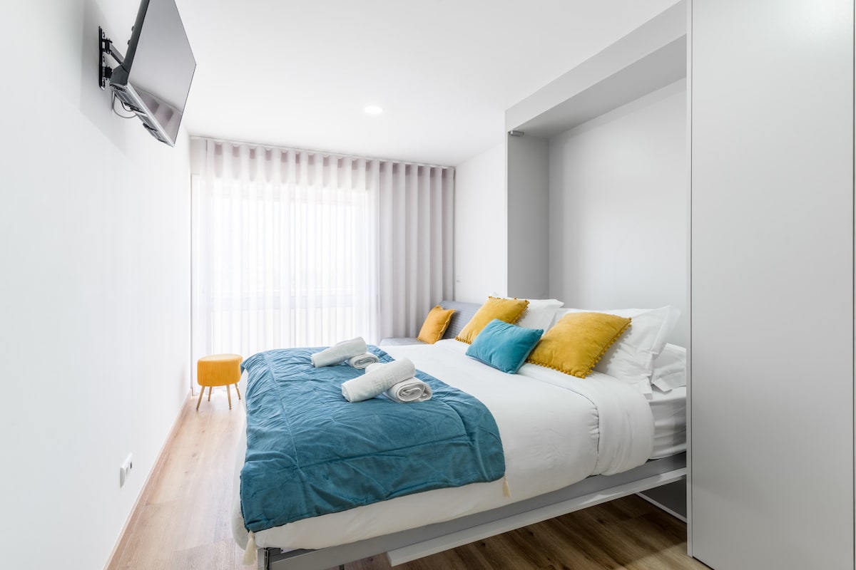 A compact sleeping area features a built-in double bed with a light blue bedspread and decorative yellow pillows. A wall-mounted TV is present, with soft natural light filtering through sheer curtains of a large window.