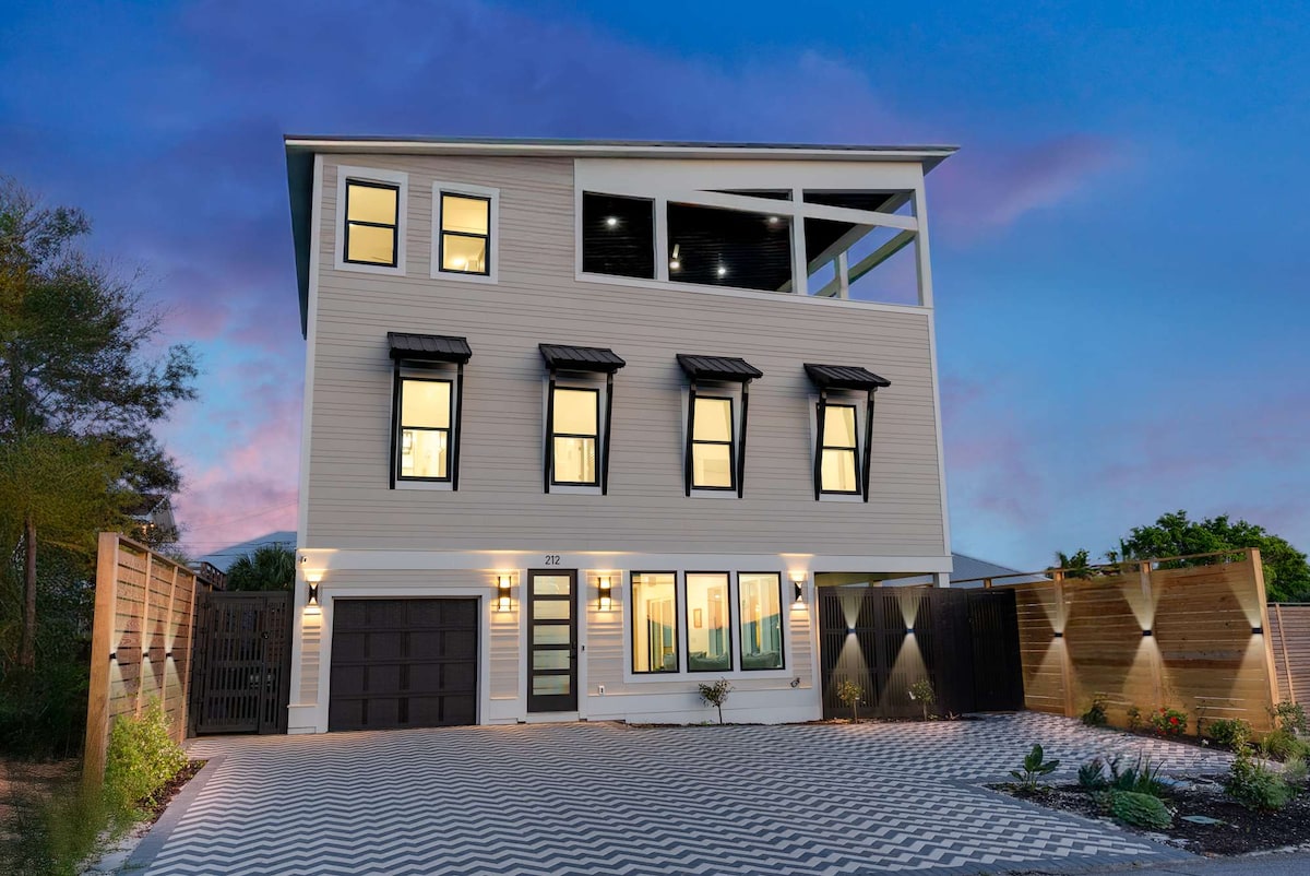 The exterior of the home showcases a contemporary three-story design with large windows framed in black. A spacious driveway is visible, featuring a chevron pattern. The rooftop deck is highlighted, offering potential views, while landscaped areas flank the entrance, enhancing the property’s inviting appearance.